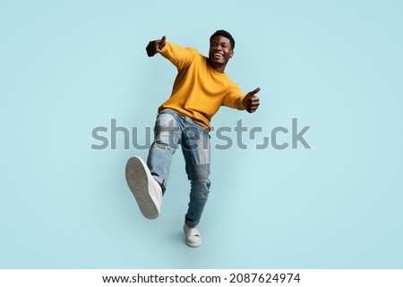 Carefree african american young man in casual outfit showing thumb ups and raising leg up, happy black guy posing on blue studio background, full length shot, panorama with copy space