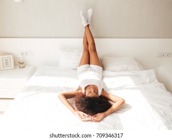 Carefree African Woman In Pajamas Lying In Bed With Raised Legs