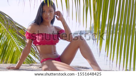 Carefree Asian female in pink bikini sitting on rock at tropical coastline and looking at camera among green leaf of palm at Thailand