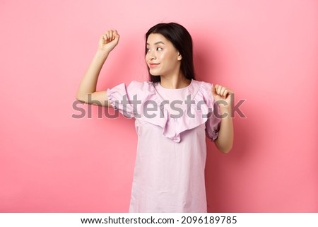 Carefree asian girl dancing, raising hand up and looking left at logo, standing in romantic dress against pink background