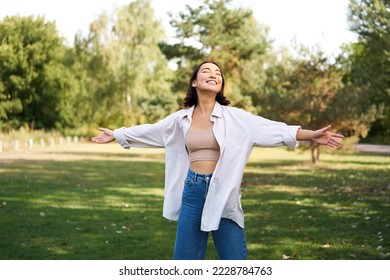 Carefree asian girl laughing and dancing in park, enjoying summer sunny day, raising hands up and breathing fresh air.