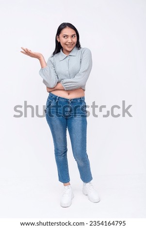 A carefree asian woman with mid-length straight hair in a long sleeved cutoff blouse and blue jeans saying oh well casually. Possible college student pointing left. Isolated on a white backdrop.