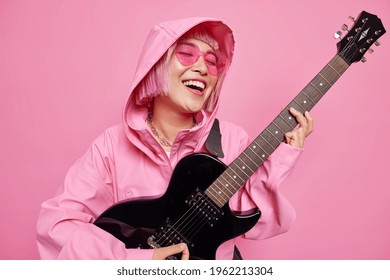 Carefree awesome hipster girl plays acoustic guitar practices before music performance dressed in jacket with hood has pink hair poses indoor. Talented female artist with musical intstrument