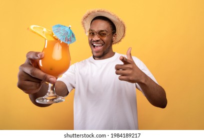 Carefree black guy in casual wear holding tropical cocktail and pointing at camera on yellow studio background. Cool young man drinking yummy alcohol-free beverage on summer vacation - Powered by Shutterstock