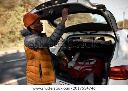 Carefree black man put backpack with equipment in cars trunk and close it. Car ride to new adventures, seasonal fun trip, joy of life. Travelling, road journey, open new horizons by yourself concept