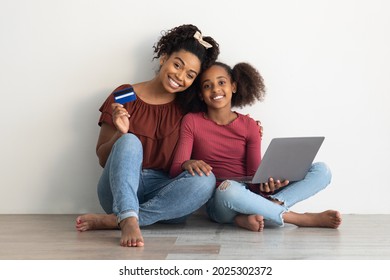 Carefree Black Young Mother And Cute Teen Daughter Shopping Online Together, Sitting On Floor Over Empty While Wall At Home, Holding Credit Card And Using Modern Laptop, Making Order On Internet