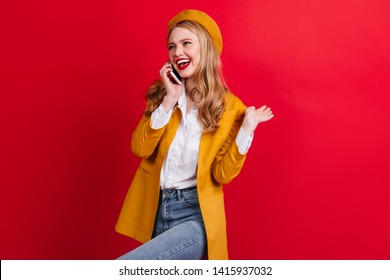 Carefree Blonde Girl Talking On Phone And Dancing. Studio Shot Of Fashionable French Woman In Beret Holding Smartphone On Red Background.