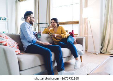 Carefree Boyfriend And Girlfriend Sitting On Comfortable Sofa In Sunlit Living Room While Holding Drinks And Laughing At Inside Jokes