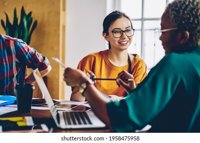 Carefree Caucasian employee in optical spectacles for provide eyes correction talking with dark skinned colleague discussing paperwork during collaborative briefing at desktop, business concept