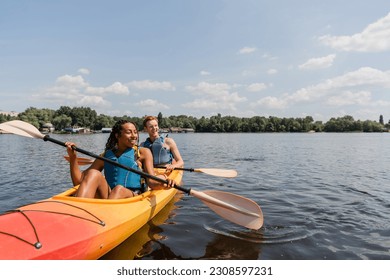 carefree and charming african american woman and young redhead man in life vests sailing in sportive kayak on calm water surface under blue sky with clouds on summer day - Powered by Shutterstock