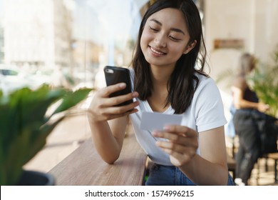 Carefree Cheerful Asian Woman In Urban Cafe, Sit Near Window At Coffee Table, Hold Smartphone, Credit Card, Entder Digits As Send Money Friend, Sharing Bill With Coworker, Smiling As Shopping Online