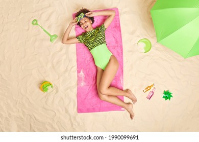 Carefree Cheerful Slim Woman Dressed In Bikini Wears Snorkeling Mask Lies On Pink Towel At White Sand Beach Enjoys Awesome Summer Travel Laughs Happily Looks From Above. Relaxation And Vacation