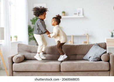 Carefree childhood. Happy energetic african american children jumping on sofa while playing game together at home, small active kids brother and sister having fun in living room
