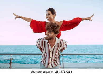 Carefree couple having fun with a piggyback ride by the sea, woman spreading her arms like a bird, enjoying freedom and happiness. - Powered by Shutterstock