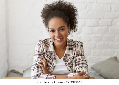 Carefree dark-skinned woman with curly hairstyle sitting crossed hands at table looking with her dark charming eyes and gentle smile into camera. Young mixed race lady waiting for her lover at cafe