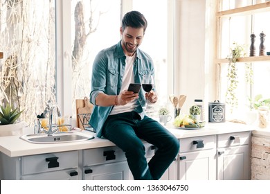 Carefree Day. Handsome Young Man In Casual Wear Drinking Wine And Using His Smart Phone While Sitting In The Kitchen At Home