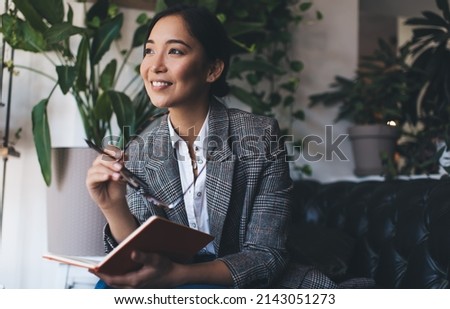 Carefree female with eyeglasses and education textbook for studying and learning smiling while dreaming about knowledge results in university college, happy Asian journalist with notepad planning