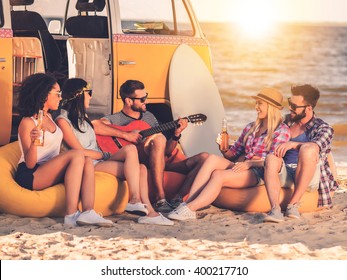 Carefree fun. Group of joyful young people drinking beer and playing guitar while sitting on the beach near their retro minivan - Powered by Shutterstock