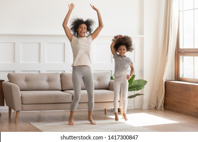 Carefree funny african family young mom having fun with cute little kid girl jump dancing in living room, happy mixed race mother with small child daughter laugh do morning exercise together at home