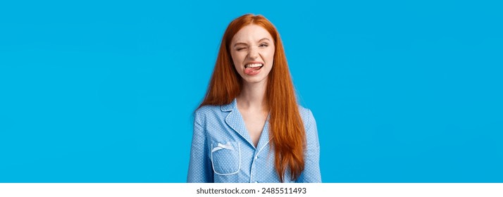 Carefree and funny pretty redhead teenage girl dont want sleep during sleepover girlfriends, showing tongue and fool around, making fun grimaces, winking joyfully, wear nightwear, blue background.