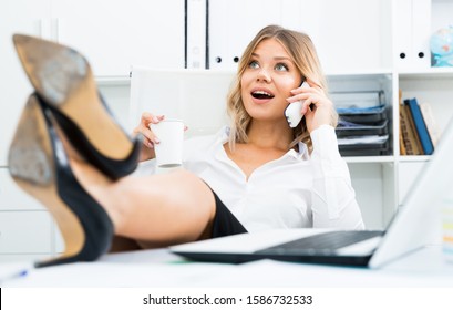 Carefree girl in business clothes is putting her feet on table and talking on phone