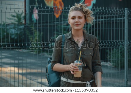 Carefree girl holding a big glass with a cold drink while tasting, drinking, enjoying it on an urban background, making funny face expressions.