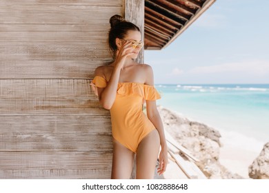 Carefree Girl In Vintage Swimwear Standing Near Wooden House And Looking At Sea. Outdoor Photo Of Gorgeous Brunette Woman With Trendy Hairstyle Relaxing At Resort.