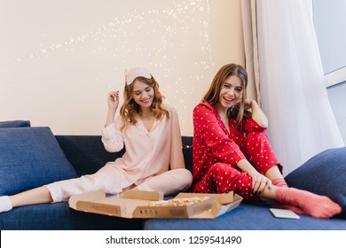 Carefree Girls In Cute Night-suits Eating Pizza Together. Romantic Young Woman In Red Pajama Sitting On Sofa With Sister And Enjoying Fast Food.