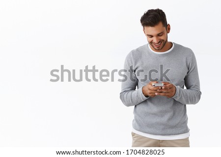 Carefree good-looking boyfriend staying touch with girlfriend abroad watching funny video-message in smartphone app, holding mobile phone gazing satisfied display, standing white background