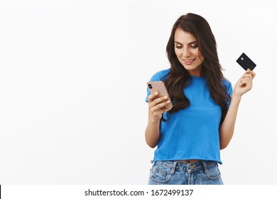 Carefree Good-looking Female In Blue T-shirt, Casually Using Smartphone, Holding Credit Card, Send Money Via Bank Account Using Application, Enter Bill Details To Make Purchase Online