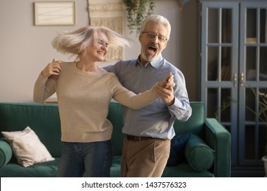 Carefree Happy Active Old Senior Couple Dancing Jumping Laughing In Living Room, Cheerful Retired Elder Husband Holding Hand Of Mature Middle Aged Wife Enjoy Fun Leisure Retirement Lifestyle At Home