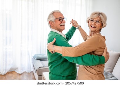 Carefree Happy Active Old Senior Couple Dancing Jumping Laughing In Living Room, Cheerful Retired Elder Husband Holding Hand Of Mature Middle Aged Wife Enjoy Fun Leisure Retirement Lifestyle At Home