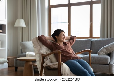 Carefree happy millennial indian woman resting in cozy armchair with folded arms behind head, enjoying peaceful calm stress free leisure weekend time alone in modern living room, breathing fresh air.