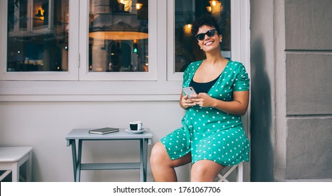Carefree happy overweight woman in sunglasses and green dress using mobile phone during recreation in cafe patio looking at camera