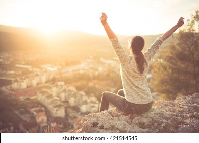 Carefree happy woman sitting on top of mountain edge cliff enjoying sun on her face raising hands in sunlight rays.Enjoying nature sunset.Freedom.Enjoyment.Relaxing in mountains at sunrise.Daydreaming