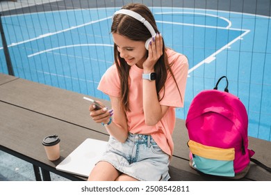 Carefree high school girl teenager pupil college student in casual clothes with wireless headphone holding using mobile phone listening enjoying music song sitting in sport court yard - Powered by Shutterstock