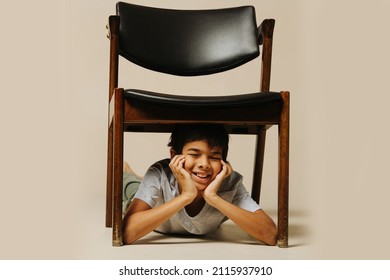 Carefree Indian Boy Playing With An Armchair Lying Under It With Eyes Closed, Leaning On His Hands. Over Beige Background.