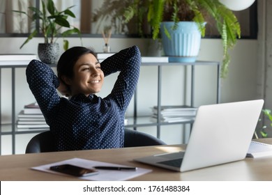Carefree indian employee resting after busy fruitful workday leaned on office chair puts hands behind head feels satisfied by work done, achievements, job promotion, looking in distance out the window