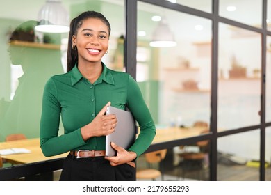 Carefree inspired african-american female employee standing in modern office space and holding laptop, cheerful black businesswoman in green shirt looks at the camera with light friendly smile