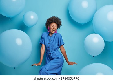 Carefree joyful curly woman dances happily, dressed in blue dress, chills at party around inflated helium balloons, feels playful, enjoys favorite holiday, has upbeat festive mood. Moment of joy - Powered by Shutterstock