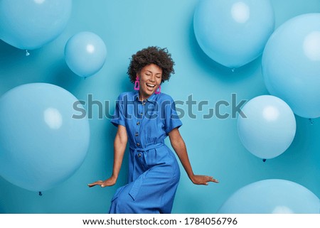 Carefree joyful curly woman dances happily, dressed in blue dress, chills at party around inflated helium balloons, feels playful, enjoys favorite holiday, has upbeat festive mood. Moment of joy
