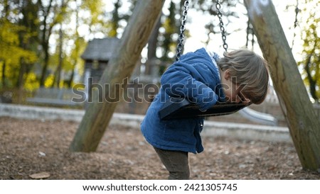 Carefree little boy leaning on park swing during autumn day wearing blue jacket, child twisting and turning into swing playing by himself during fall season outdoors