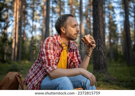 Carefree man eating sandwich snack during hike walk travel camping vacation, feels calm relax in nature landscape. Happy hiking male resting taking break with healthy food in forest. Autumn nature.