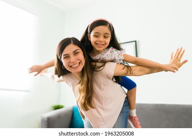 Carefree Mom And Little Girl Laughing While Playing At Home. Hispanic Beautiful Mother Carrying Her Daughter On The Back Like An Airplane