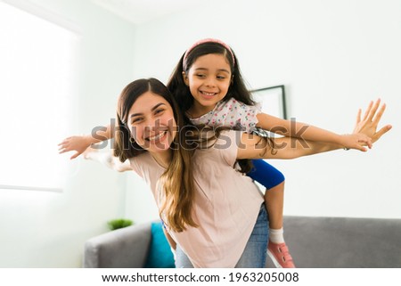 Carefree mom and little girl laughing while playing at home. Hispanic beautiful mother carrying her daughter on the back like an airplane