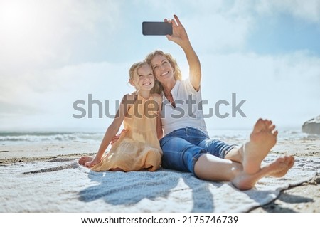 Carefree mother and daughter taking a selfie while sitting on the beach. Happy little girl and grandmother smiling while taking a picture on a cellphone while on holiday. Mom and daughter bonding
