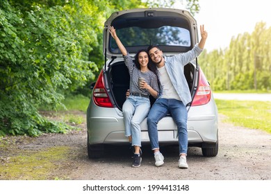 Carefree multiracial millennial couple having break at countryside while car trip, sitting on open trunk, drinking coffee, embracing and cheerfully smiling at camera, raising hands up, free space
