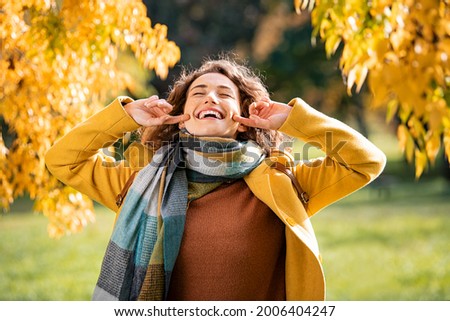 Carefree natural beauty woman enjoying freedom with yellow leaves on trees in background. Playful girl wearing coat and scarf laughing during autumn outdoor. Beautiful girl using finger while laughing