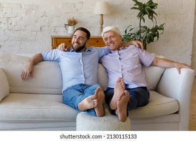Carefree older man his grown up son embracing look into distance relax together on sofa put their feet on footstool, daydreaming together at comfort home. Unity, multigenerational family bond concept