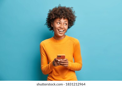Carefree pleased female student uses mobile phone for online distant education looks gladfully aside and smiles broadly browses through internet during free time checks mailbox poses indoor.